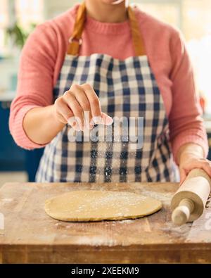 Primo piano di una donna a casa in cucina che sparge la farina sull'impasto arrotolato sul piano di lavoro o sul bancone Foto Stock