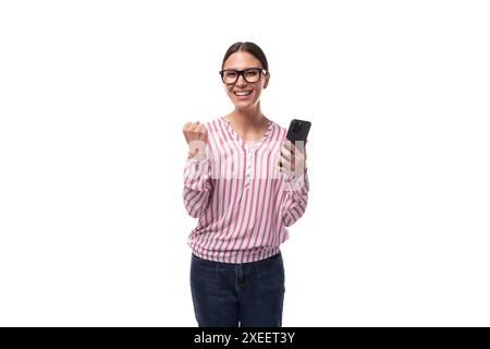La giovane segretaria con gli occhiali si rallegra di tenere uno smartphone tra le mani su uno sfondo bianco Foto Stock