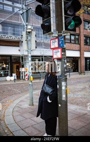 Una ragazza con un cappotto nero e una borsa a mano a un semaforo all'incrocio di una città tedesca in un giorno d'autunno. Una giovane donna sta aspettando un tr verde Foto Stock