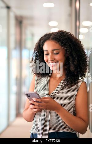 Donna d'affari in piedi nel corridoio del moderno edificio per uffici utilizzando il telefono cellulare Foto Stock