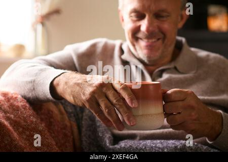 Primo piano dell'uomo maturo a casa in inverno salta sul divano con una bevanda calda calda calda calda di caffè in tazza Foto Stock