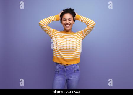 Giovane donna latina riccia bruna allegra e carina in maglione a righe giallo Foto Stock