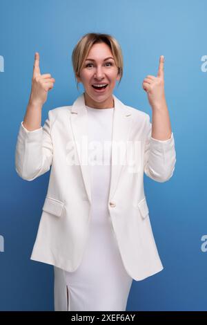 Allegra e bella bionda, giovane donna europea vestita con una giacca bianca e il vestito punta le dita sulla pubblicità Foto Stock