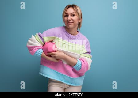 Foto ravvicinata di un'attraente donna bionda in un maglione a righe con un salvadanaio su sfondo blu con copia spa Foto Stock