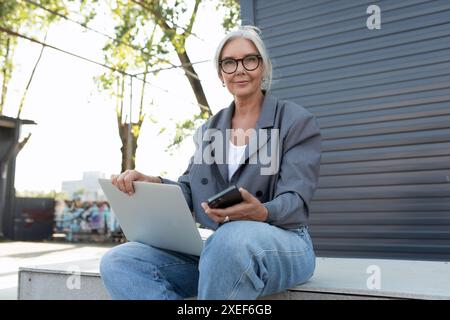 Una bella donna d'affari dai capelli grigi, vestita con un'elegante giacca grigia e jeans seduti su una panchina con un laptop Foto Stock