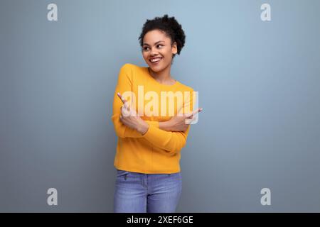 Giovane donna latina intelligente con capelli afro che punta il dito verso il lato su sfondo grigio con spazio di copia Foto Stock