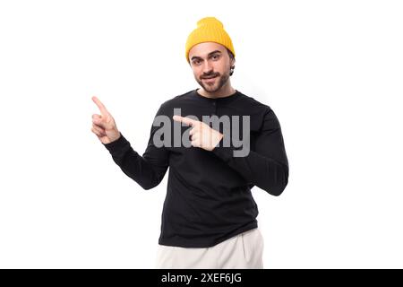 Un giovane ragazzo di 30 anni, brutale e bello, vestito con una giacca nera e un cappello giallo, punta al muro con la mano Foto Stock