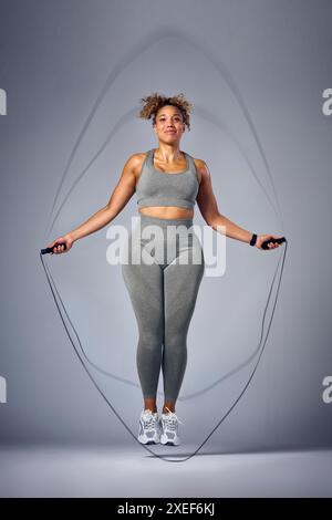 Studio Portrait Shot of Woman in Gym fitness Clothing Skipping with Rope on Grey background Foto Stock