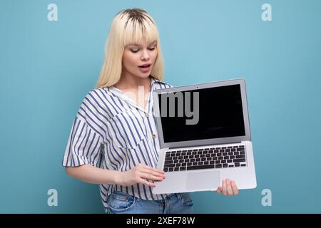 La bella ragazza bionda in un outfit casual mostra lo schermo di un notebook con un mockup Foto Stock