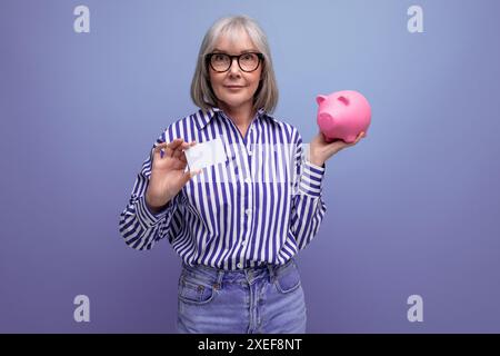 Risparmi sulle pensioni. una donna di mezza età con i capelli grigi tiene un salvadanaio con soldi su uno sfondo luminoso dello studio Foto Stock