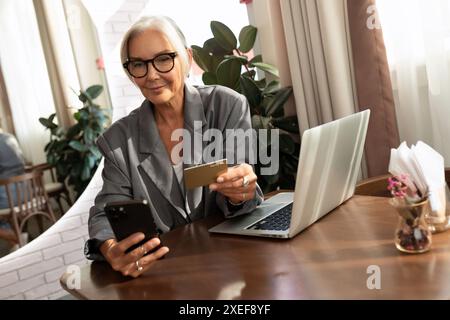 Una donna d'affari europea dai capelli grigi si siede in un bar e tiene in mano una carta di credito e uno smartphone Foto Stock