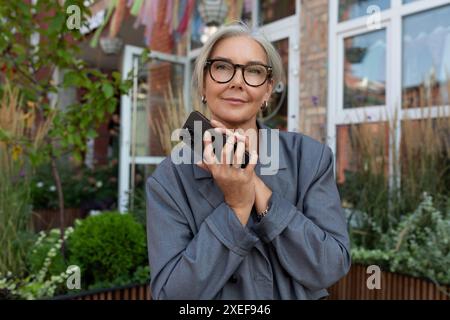 Una donna anziana ben curata con capelli e occhiali grigi è vestita con una giacca e un jeans grigi, tenendo in mano uno smartphone a. Foto Stock