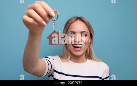 Giovane donna felice e felice che tiene la casa portachiavi e si rallegra di acquistare immobili su sfondo blu isolato Foto Stock