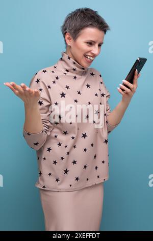 Gioiosa giovane donna caucasica con un taglio di capelli corto vestito con un abito beige guarda un video al telefono Foto Stock
