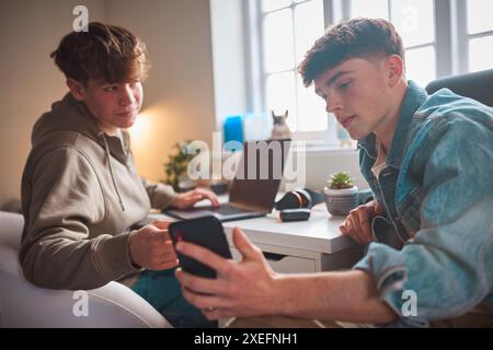 Due ragazzi adolescenti a casa in camera da letto che guardano messaggio o social media sul telefono cellulare e ridono Foto Stock