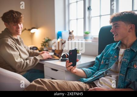 Due ragazzi adolescenti a casa in camera da letto che guardano messaggio o social media sul telefono cellulare e ridono Foto Stock