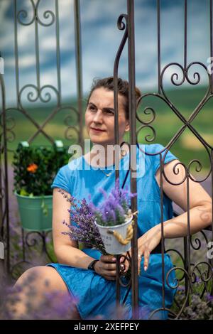 Una donna con un abito blu si siede con cura, tenendo in mano un bouquet di lavanda. È circondata da una recinzione in ferro battuto con vasi appesi. Foto Stock