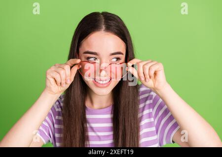 Foto di un bel look da donna in uno spazio vuoto togliete gli occhiali da sole dal cuore vestito con abito viola a righe isolato su sfondo di colore verde Foto Stock