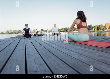 Lezione di yoga all'aperto con istruttore vicino al fiume. Le giovani donne siedono a gambe incrociate sui tappetini e meditano su un molo di legno. Foto Stock