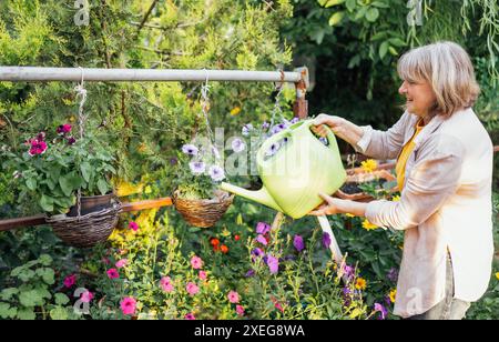 Un'anziana donna sorridente annaffia i fiori in vasi appesi da una lattina da innaffiare. Si prende cura di una donna anziana positiva in abiti casual Foto Stock