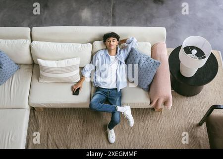 Rilassatevi sul divano, un giovane che tiene lo smartphone e guarda la fotocamera Foto Stock