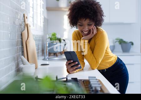 Donna sorridente che usa lo smartphone in una cucina moderna e si diverte a casa Foto Stock