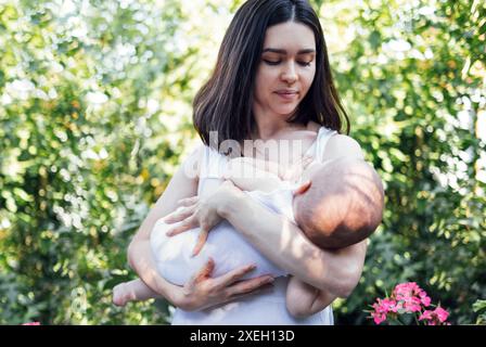 Una bella madre in abiti bianchi sta dondolando il suo bambino tra le braccia in giardino. Foto Stock