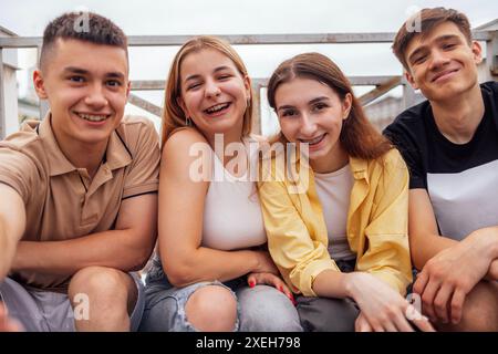 Quattro simpatici adolescenti che si fanno un selfie. Amici divertenti scattano una foto di sé stessi al telefono all'aperto. Ragazzi e ragazze sorridenti seduti Foto Stock