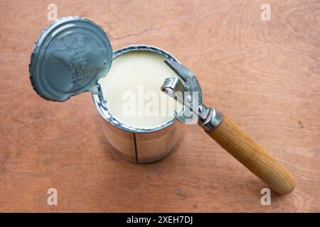 Lattina metallica aperta di latte condensato e apriscatole in metallo su sfondo marrone in legno. Foto Stock