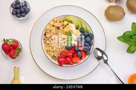 Granola con fragole, kiwi, banana e mirtilli in un piatto rotondo su un tavolo bianco. Foto Stock