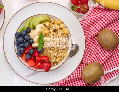 Granola con fragole, kiwi, banana e mirtilli in un piatto rotondo su un tavolo bianco. Foto Stock