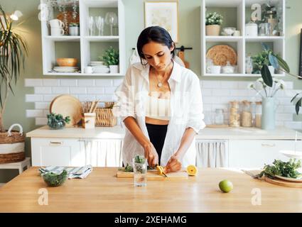 Una giovane e attraente donna prepara una deliziosa limonata fresca e taglia il limone e la menta su un piedistallo di legno. Un'affascinante bruna vegana Foto Stock