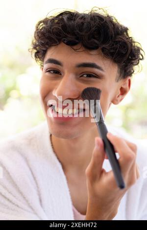 Giovane sorridente che applica il trucco con la spazzola, indossando l'abito bianco a casa Foto Stock