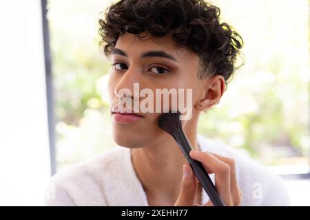 Applicazione del trucco, giovane uomo che usa il pennello per rifinire i contorni a casa, concentrandosi sul viso Foto Stock