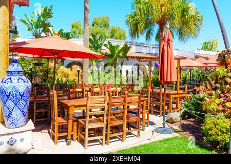 San Diego, California - 16 aprile 2024: Tavoli e sedie in legno disposti in un'area pranzo all'aperto con ombrelli rossi, circondati da una lussureggiante vegetazione Foto Stock