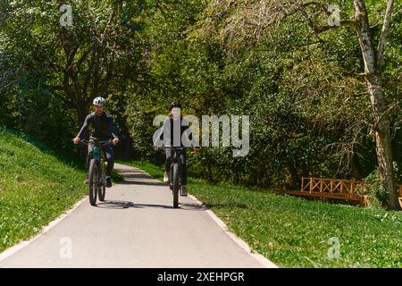 Una coppia beata, adornata di attrezzatura da ciclismo professionale, gode di una romantica corsa in bicicletta attraverso un parco, circondato dalla moderna nat Foto Stock