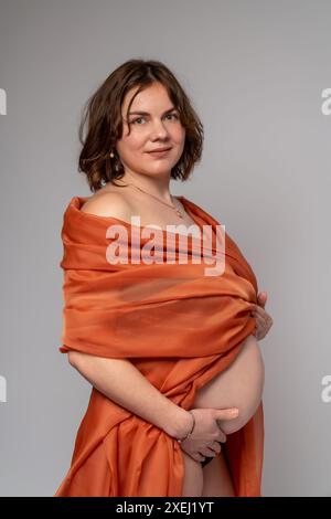 Una donna incinta in un mantello arancione tiene le mani sullo stomaco su uno sfondo grigio chiaro. Gravidanza, maternità, preparazione e aspettativa di un Foto Stock