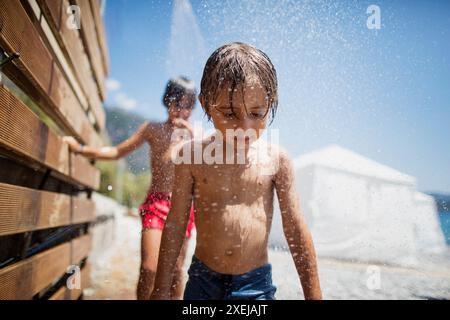 Un bambino si gode una doccia rinfrescante su una spiaggia soleggiata, mentre un altro ragazzo cammina sullo sfondo. Foto Stock