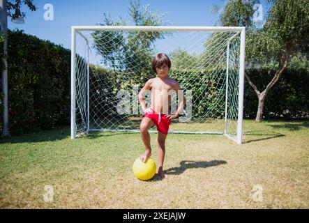 Un ragazzo si erge con il piede su un pallone da calcio davanti a una porta, trascorrendo una giornata estiva sul campo da calcio. Foto Stock