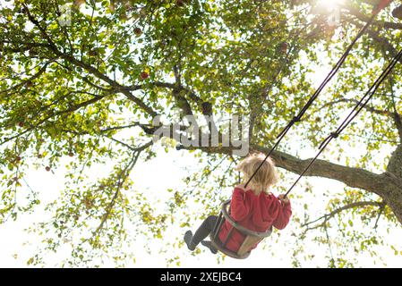 Bambino che oscilla sotto un albero con la luce del sole che filtra attraverso le foglie Foto Stock