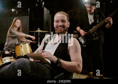 Ritratto medio di un giovane musicista rock caucasico seduto su una sedia con blocco note in mano sorridente alla macchina fotografica Foto Stock
