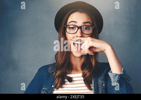 Donna sorpresa, ritratto e volto divertente con baffi alle dita, cappello o espressione goffa su sfondo grigio studio. Donna, hipster o comica Foto Stock