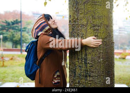 La giovane donna amante della natura abbraccia l'albero e lo guarda e sorride, indossa una felpa con cappuccio marrone, si trova nel parco a toccare la corteccia con le mani A. Foto Stock