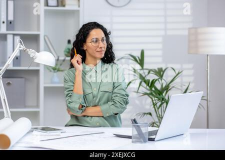 Architetto seduto alla scrivania in un ufficio moderno, tenendo una matita e guardando premuroso. Pianificazione e progettazione di progetti utilizzando notebook, progetti e tablet digitali. Area di lavoro luminosa e organizzata Foto Stock