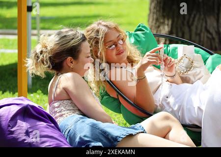 Due ragazze sorridenti che guardano lo schermo dello smartphone sedute in borse di fagioli nel parco cittadino in estate Foto Stock