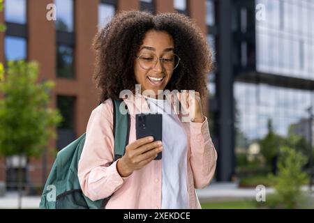 Giovane studentessa che indossa occhiali e zaino per celebrare le buone notizie mentre guarda uno smartphone vicino al campus universitario. Ambiente esterno con edificio moderno sullo sfondo. Foto Stock