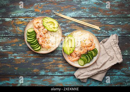 Due ciotole di ceramica bianca con riso, gamberetti, avocado, verdure, semi di sesamo e bacchette su un colorato sfondo rustico in legno con vista dall'alto Foto Stock