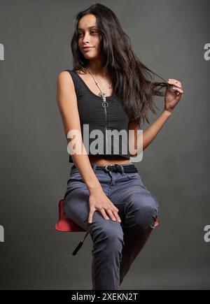 La modella ispanica adolescente è seduta su una sedia, la donna guarda di profilo mostrando i suoi bellissimi capelli neri. È vestita alla moda con una t nera Foto Stock