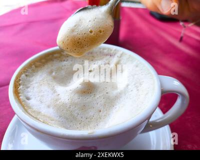 Torbole, Lago di Garda, Italia - 28 giugno 2024: Cappuccino in un ristorante con crema di latte o crema di latte su un cucchiaio, caffè di concetto - il cappuccino perfetto con crema di caffè e schiuma di caffè. *** Cappuccino in einem ristorante mit aufgeschäumter Schaum aus Milch, bzw. Milchschaum am Löffel, Konzept Kaffee - der perfekte Cappuccino mit Kaffee crema und Kaffeeschaum. Foto Stock