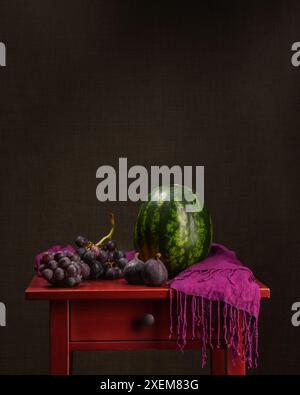 Natura morta con il cibo. Anguria, uva, fico su un tavolo di legno rosso con scialle viola. Sfondo scuro. Colori autunnali. Cartolina calma. calendario.vegano Foto Stock